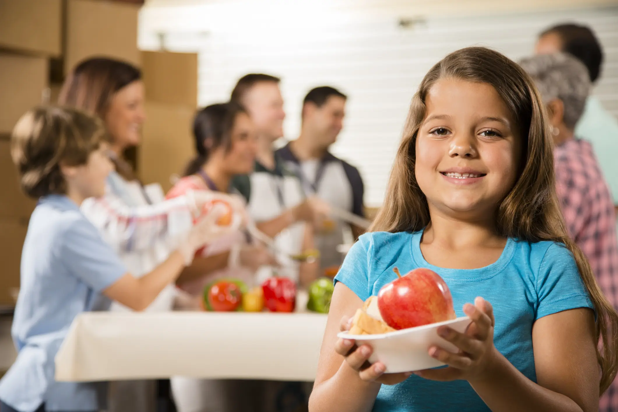 Multi-ethnic and mixed-age group of volunteers serve food to needy people at a community "soup kitchen" or food pantry location. A hungry little girl is in the foreground holding a bowl of food she just received. The volunteers wear aprons, lanyards, and gloves. Vegetable, fruit, and soup food items are on the table with boxes all around the group.  Service, kindness, charity, community outreach themes.