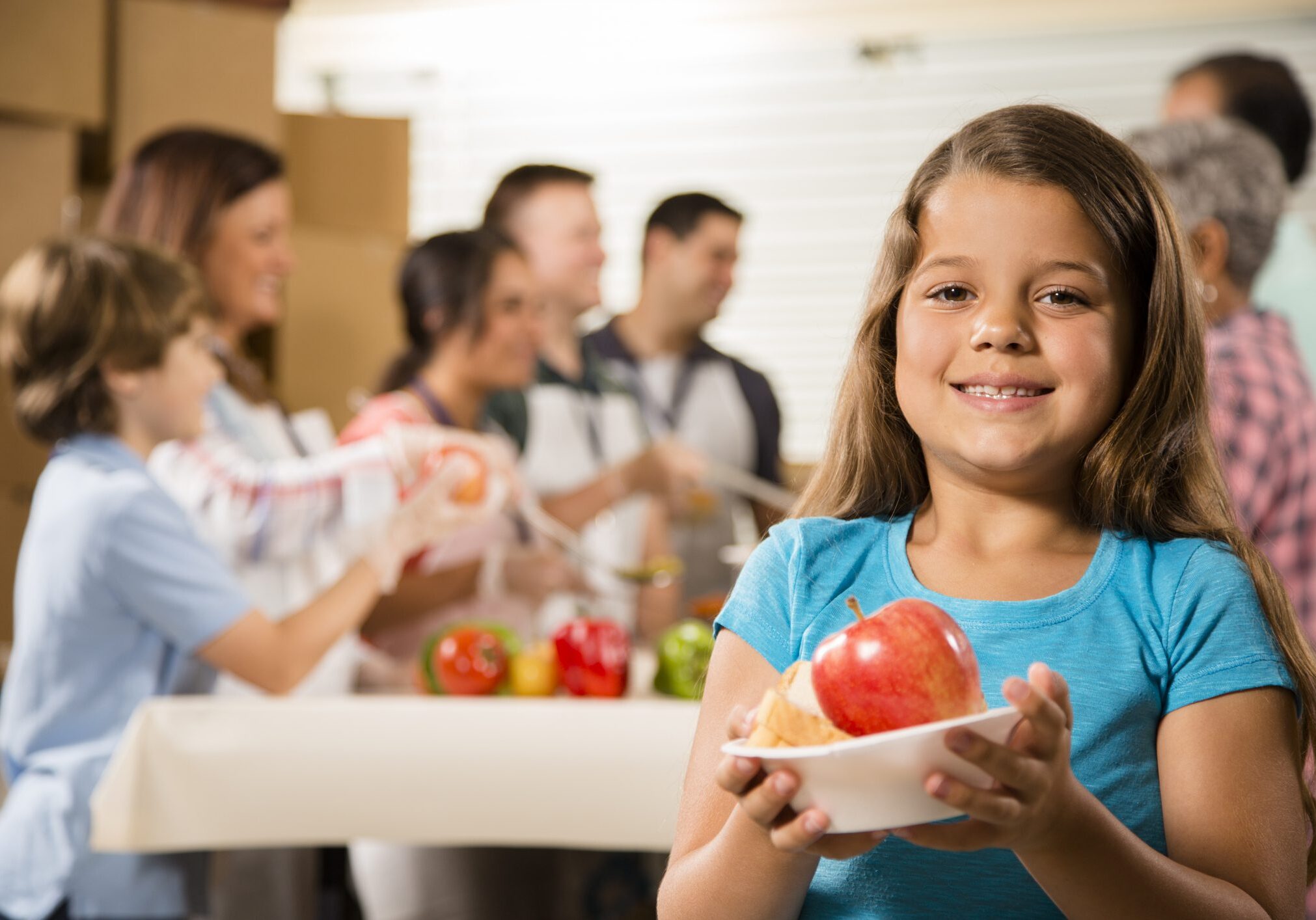 Multi-ethnic and mixed-age group of volunteers serve food to needy people at a community "soup kitchen" or food pantry location. A hungry little girl is in the foreground holding a bowl of food she just received. The volunteers wear aprons, lanyards, and gloves. Vegetable, fruit, and soup food items are on the table with boxes all around the group.  Service, kindness, charity, community outreach themes.